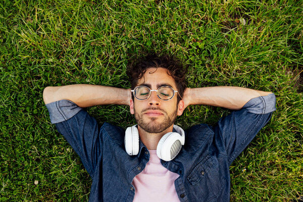 Outdoor portrait of a handsome young hipster man lying in the garden with headphones.