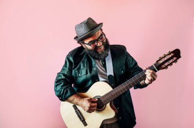 Mutlu sakallı adam pembe arka planda akustik gitar çalıyor..