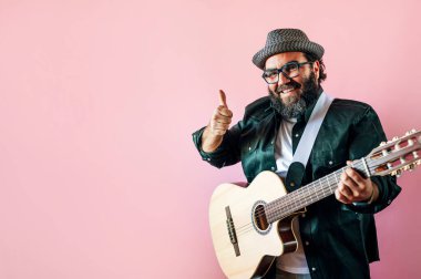 Mutlu sakallı adam pembe arka planda akustik gitar çalıyor ve baş parmağını kaldırıyor..