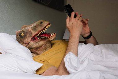 Kertenkele maskeli bir adam otel yatağında akıllı telefon kullanıyor..