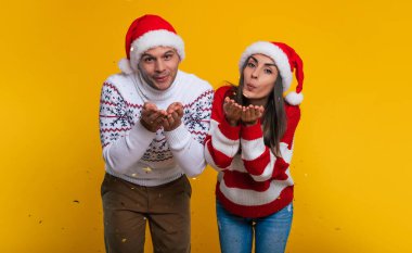 Sarı arka planda izole bir şekilde eğlenirken, Noel Baba şapkaları ve süveterleriyle birbirine aşık genç ve mutlu çift.