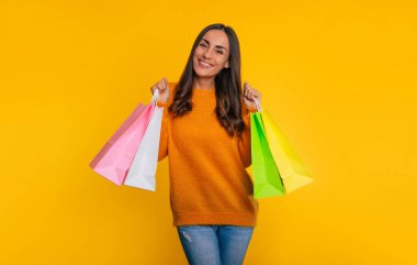 Şirin, şık, genç, gülümseyen bir kadın, sarı arka planda izole edilmiş bir halde dururken bir sürü renkli alışveriş poşetini elinde tutuyor.