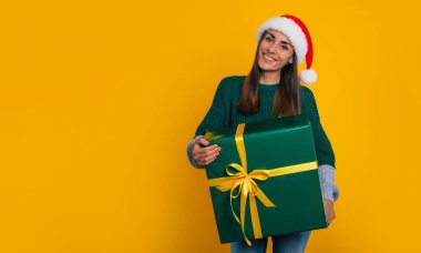 Noel Baba şapkalı güzel, mutlu bir kadın elinde hediye kutusuyla sarı arka planda yalnız poz veriyor.