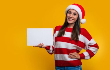 Modern, büyüleyici, güler yüzlü bir kadının, Noel tarzı giysiler içinde beyaz tahtayı elinde tutarken ve kameraya bakarken çekilmiş bir fotoğrafını çek.
