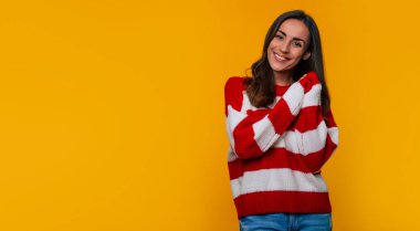 O poz verirken, kameraya bakarken ve sarı arka planda izole edilirken, güzel, mutlu, gülümseyen, çizgili süveterli bir kadın.
