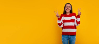 Mutlu gülümseyen genç, güzel, modern kadın sarı arka planda izole bir şekilde poz verirken zafer işareti gösteriyor.