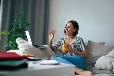Dizüstü bilgisayarda çalışan ve büyük koltukta oturup evinde kahve içerken internette sohbet eden tatlı, serbest çalışan bir kadın.