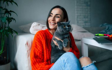 Güzel, güzel, esmer bir kadın, sevimli İngiliz kedisini kucağına alıp kucaklıyor ve evinde yerde dinleniyor.