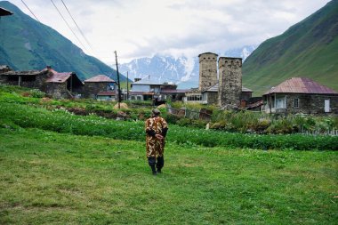 Ushguli, Georgia - August 2015: Georgian old woman walking in Ushguli village with rock tower houses in Svaneti, Georgia. These are typical Svaneti defensive tower houses found throughout the village clipart