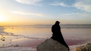 Şiraz, İran - Mayıs 2019: Maharloo Pembe Gölü kıyısındaki bir kayanın üzerinde oturan tesettürlü İranlı siyah elbiseli kadın