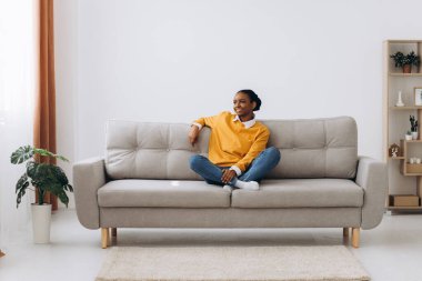 Genç siyahi bayan hafta sonunun tadını çıkarıyor ve evdeki rahat kanepede dinleniyor. Afro-Amerikalı bir kadın oturma odasında kanepede oturuyor, kollarını açıp gülümsüyor ve tedirgin ve mutlu görünüyor.