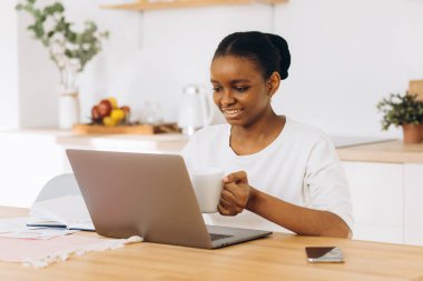 Genç siyah kadın mutfakta oturup kahve içiyor ve laptopla evden çalışıyor..
