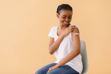 Genç Afrikalı Amerikalı kadın ilk aşıdan memnun, yara bandı takıyor, kolunu kaldırıyor ve arkadaşlarına iğneden bahsediyor. Bej arkaplanda izole edilmiş.