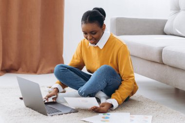 Genç, güzel siyah bir üniversite öğrencisi yerde oturuyor, kahve içiyor ve dizüstü bilgisayarında proje yapıyor.