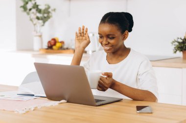 Genç siyah kadın mutfakta oturup kahve içiyor ve bilgisayardan video görüşmesi yapıyor..