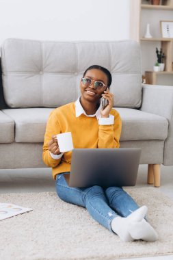 Güzel siyah üniversite öğrencisi yerde oturuyor, kahve içiyor, telefonla konuşuyor ve dizüstü bilgisayarla proje yapıyor.