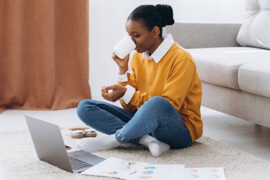 Genç, güzel siyah bir üniversite öğrencisi yerde oturuyor, kahve içiyor ve dizüstü bilgisayarında proje yapıyor.