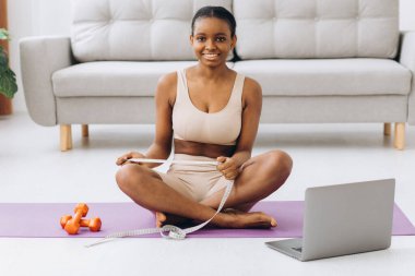 Genç siyah kadın oturma odasının arka planında belini ölçüyor. Diyet ve kilo kaybı kavramı.