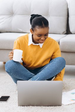 Genç, güzel siyah bir üniversite öğrencisi yerde oturuyor, kahve içiyor ve dizüstü bilgisayarında proje yapıyor.