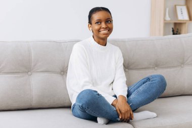 Genç siyahi bayan hafta sonunun tadını çıkarıyor ve evdeki rahat kanepede dinleniyor. Afro-Amerikalı bir kadın oturma odasında kanepede oturuyor, kollarını açıp gülümsüyor ve tedirgin ve mutlu görünüyor.