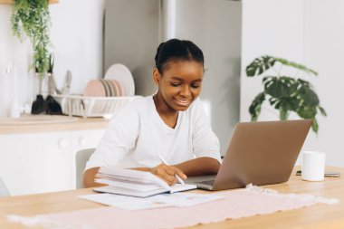 Genç siyah kadın mutfakta oturuyor ve bilgisayarla evden çalışıyor..