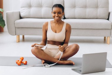 Genç siyah kadın oturma odasının arka planında belini ölçüyor. Diyet ve kilo kaybı kavramı.