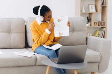 Genç bir Afro-Amerikalı kadın video görüşmesinde konuşuyor ve evdeki oturma odasında kağıt üzerinde bir şeyler gösteriyor, evden uzaklık öğrenme kavramı.