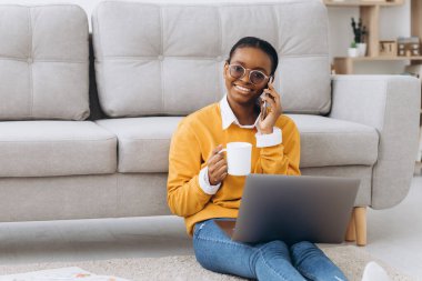 Güzel siyah üniversite öğrencisi yerde oturuyor, kahve içiyor, telefonla konuşuyor ve dizüstü bilgisayarla proje yapıyor.