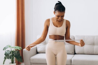 Genç siyah kadın oturma odasının arka planında belini ölçüyor. Diyet ve kilo kaybı kavramı.