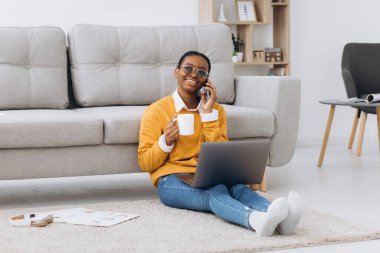 Güzel siyah üniversite öğrencisi yerde oturuyor, kahve içiyor, telefonla konuşuyor ve dizüstü bilgisayarla proje yapıyor.