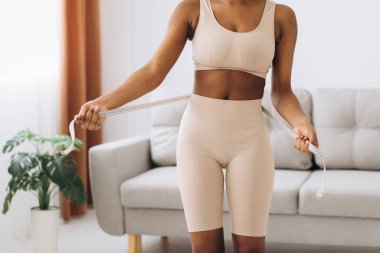 Genç siyah kadın oturma odasının arka planında belini ölçüyor. Diyet ve kilo kaybı kavramı.