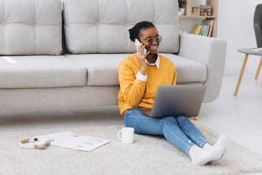 Güzel siyah üniversite öğrencisi yerde oturuyor, kahve içiyor, telefonla konuşuyor ve dizüstü bilgisayarla proje yapıyor.