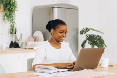 Genç siyah kadın mutfakta oturuyor ve bilgisayarla evden çalışıyor..