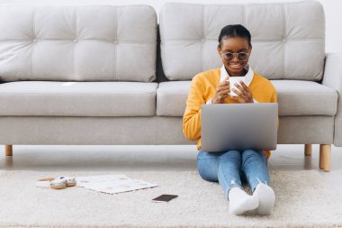 Genç, güzel siyah bir üniversite öğrencisi yerde oturuyor, kahve içiyor ve dizüstü bilgisayarında proje yapıyor.