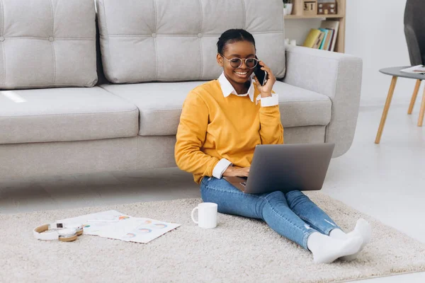 Güzel siyah üniversite öğrencisi yerde oturuyor, kahve içiyor, telefonla konuşuyor ve dizüstü bilgisayarla proje yapıyor.