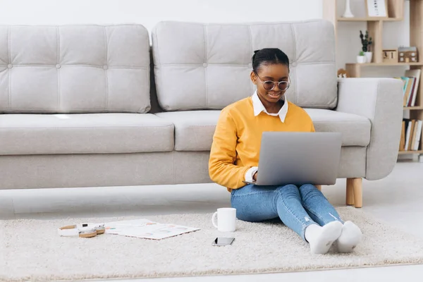 Genç, güzel siyah bir üniversite öğrencisi yerde oturuyor, kahve içiyor ve dizüstü bilgisayarında proje yapıyor.