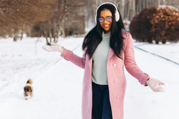 一位戴着眼镜和温暖手套的年轻的非洲裔美国妇女在一个冬季公园遛狗 — 图库照片
