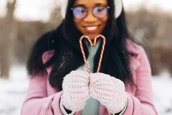 若いですアフリカ系アメリカ人女性で眼鏡と暖かいミトン保持2心形甘いキャンディー上の冬の背景にバレンタインデー — ストック写真