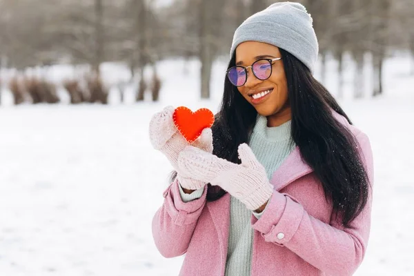 在冬天的情人节 年轻的非洲女人戴着眼镜和温暖的手套 手持一颗装饰的红心 — 图库照片