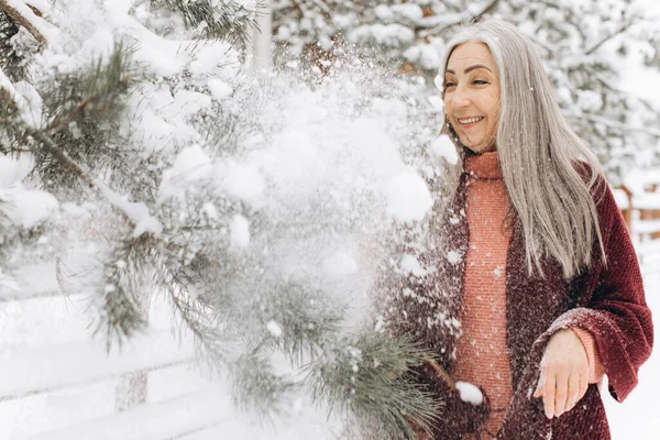 一位穿着针织毛衣和围巾 头发灰白的老妇人笑着在冬天的背景上移动了一根圣诞树树枝 — 图库照片