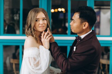 Düğün günü. Güzel Avrupalı gelin ve Asyalı damadı kafelerin ve büyük pencerelerin önünde poz veriyorlar..