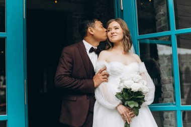 Düğün günü. Güzel Avrupalı gelin ve Asyalı damadı kafelerin ve büyük pencerelerin önünde poz veriyorlar..