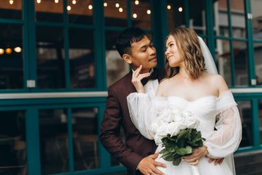 Düğün günü. Güzel Avrupalı gelin ve Asyalı damadı kafelerin ve büyük pencerelerin önünde poz veriyorlar..