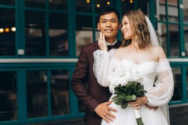 Düğün günü. Güzel Avrupalı gelin ve Asyalı damadı kafelerin ve büyük pencerelerin önünde poz veriyorlar..