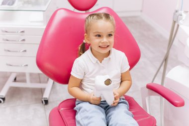 Dişçiye giderken gülümseyen küçük kız diş kliniğinde diş modelini tutuyor.