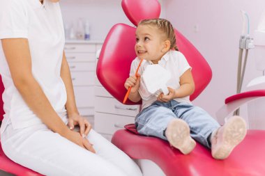 Küçük bir kız dişçi kliniğinde dişlerini nasıl fırçaladığını gösteriyor.