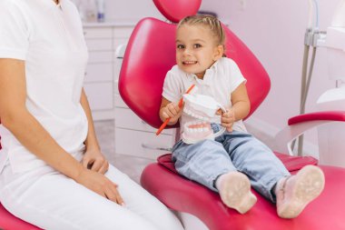 Küçük bir kız dişçi kliniğinde dişlerini nasıl fırçaladığını gösteriyor.