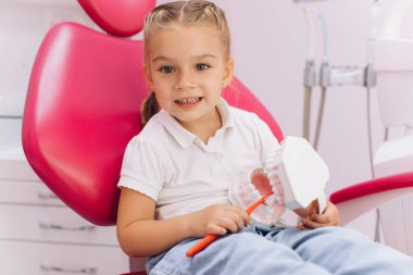Küçük bir kız dişçi kliniğinde dişlerini nasıl fırçaladığını gösteriyor.