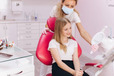 Genç kız, dişçi muayenehanesindeki bir kadın dişçiye giderken aynaya bakar.