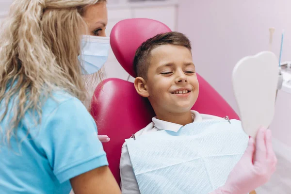 女性歯科医は鏡を持っており 歯科医院への訪問後 笑顔の男の子彼の新しい美しい笑顔を示しています — ストック写真